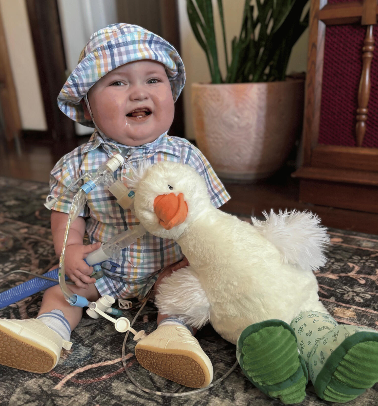 Charlie smiles a gummy smile while wearing an Easter onesie and matching hat. His fluffy Goose stuffy is sitting with him, and he holds his blue trach circuit with his right hand.